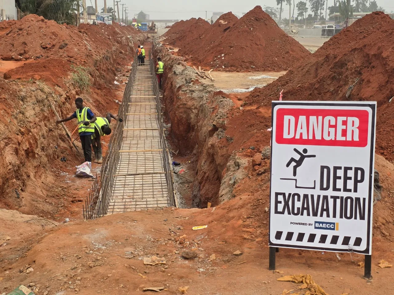 A deep drainage trench with a steel reinforcement cage laid in the base, workers in high-visibility vests, and a large deep excavation warning sign.