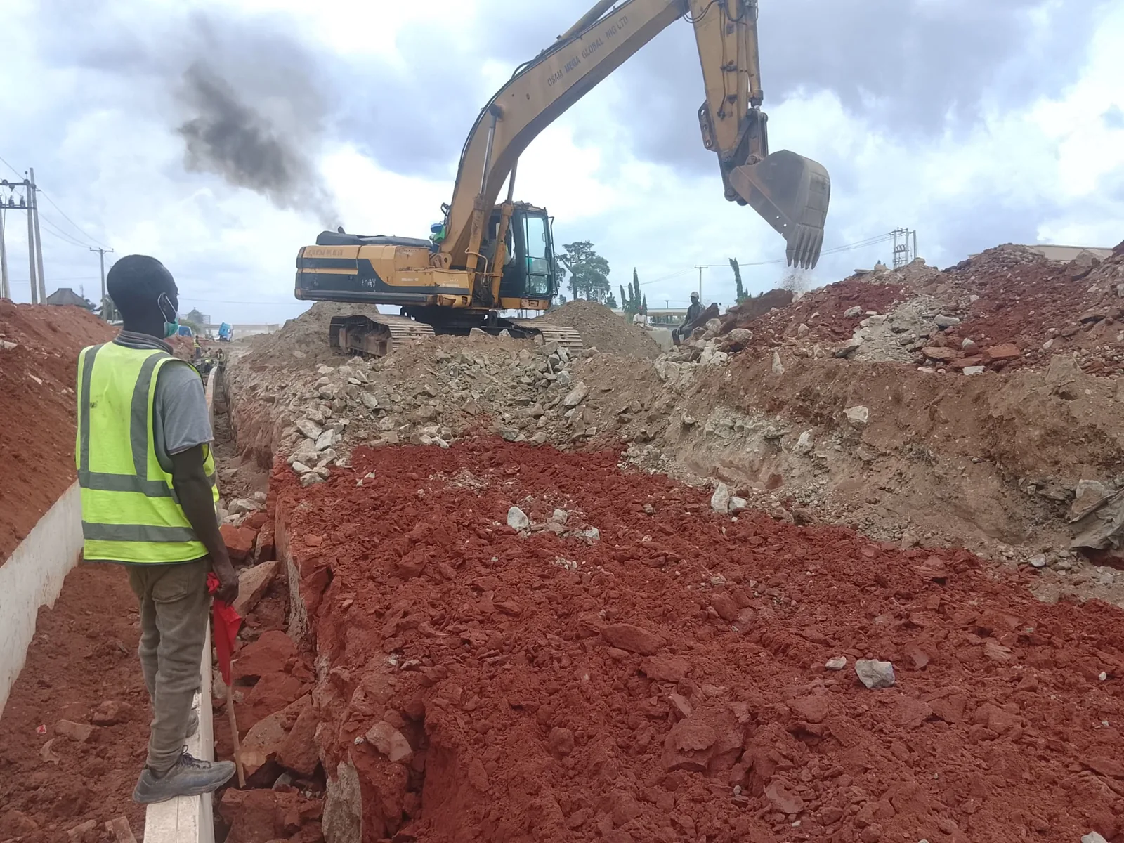 An excavator working above a drainage trench while a site worker in a high-visibility vest stands on the completed drain channel below.