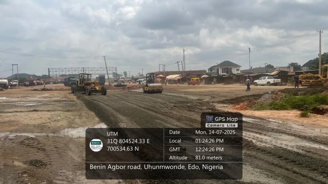 A motor grader and a roller shaping the road formation on the Benin-Agbor road, with roadside buildings alongside.
